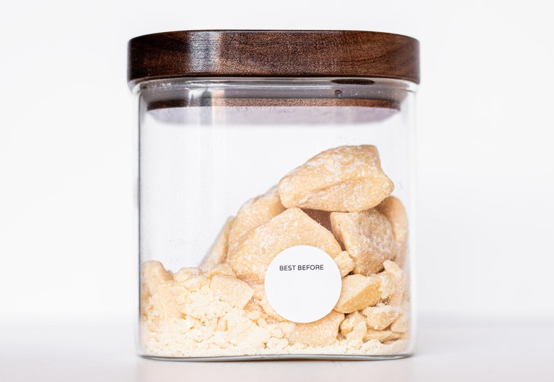May include: A glass jar with a wooden lid filled with chunks of light brown shea butter. A white label on the jar reads "BEST BEFORE".