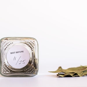May include: A clear glass jar with a white label that says "BEST BEFORE 11/25" and a pile of dried bay leaves next to it.