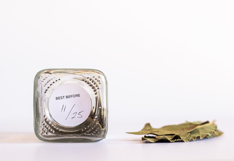 May include: A clear glass jar with a white label that says "BEST BEFORE 11/25" and a pile of dried bay leaves next to it.