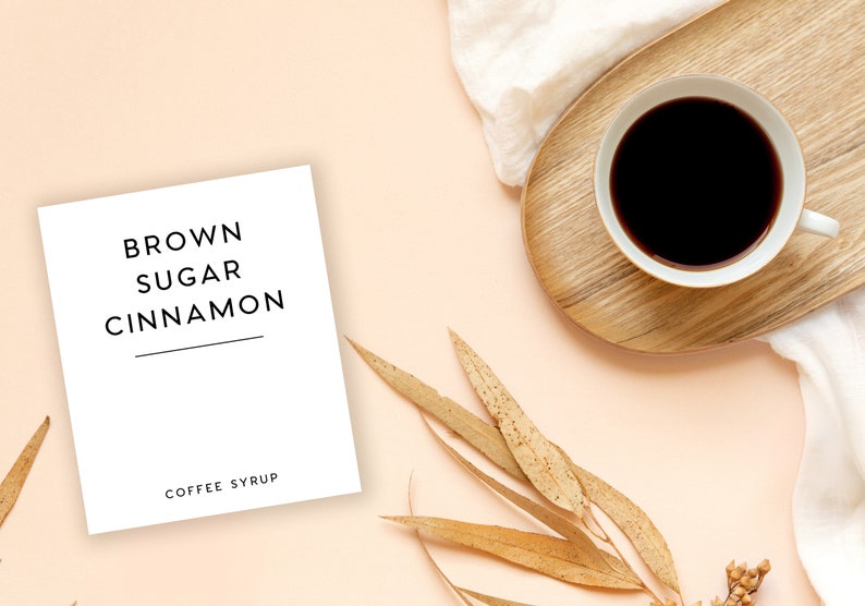 Pu&ograve; includere: Un biglietto bianco con il testo "BROWN SUGAR CINNAMON COFFEE SYRUP" su uno sfondo rosa chiaro con una tazza di caff&egrave;, foglie secche e un panno bianco.