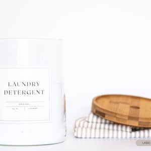 May include: A clear glass jar with a white lid and a white label that says "Laundry Detergent" with a copper scoop inside. The jar is sitting on a white surface. The label size is 4" x 4".