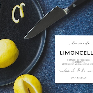 May include: A white square label with black text reading homemade LIMONCELLO, BOTTLED: OCTOBER 2023, INGREDIENTS: LEMON ZEST, VODKA, SIMPLE SYRUP, drink & be merry, DAN & KELLY, placed near two yellow lemons, a lemon peel, and a kitchen knife.