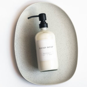 May include: A white bottle of hand soap with a black pump dispenser on a speckled gray ceramic tray. The label on the bottle reads "HAND SOAP" with the text "PREMIUM QUALITY" underneath.