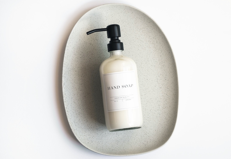 May include: A white bottle of hand soap with a black pump dispenser on a speckled gray ceramic tray. The label on the bottle reads "HAND SOAP" with the text "PREMIUM QUALITY" underneath.