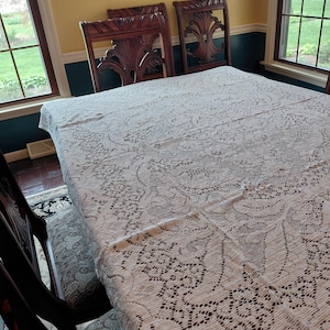 May include: A white lace tablecloth covers a rectangular table, set with ornate wooden chairs. The chairs have carved backs. The room has yellow walls, dark trim, and windows with a view of greenery.