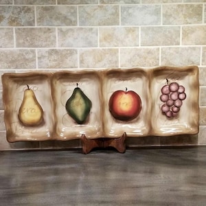 May include: A ceramic serving tray with four divided sections, each featuring a different fruit: a pear, a green pear, an apple, and grapes. The tray is decorated with a brown and white background and has a rustic, farmhouse style.