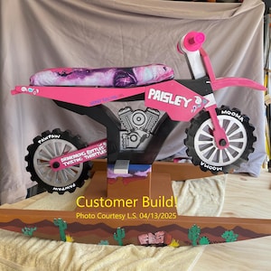 May include: A pink and black wooden rocking motorcycle with a custom paint job. The motorcycle has a purple and white patterned seat, black wheels with white lettering, and a black engine with a silver exhaust pipe. The motorcycle is on a wooden base with a desert scene painted on it. The text "PAISLEY" is written on the side of the motorcycle.