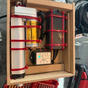 May include: A wooden enclosure containing a silver linear actuator with a yellow label, a black power adapter, and a black OONO forward and reverse relay module. The components are secured with red zip ties within the rectangular wooden frame.