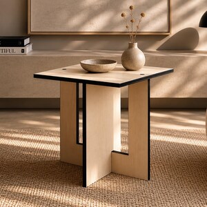 Puede incluir: Una mesa auxiliar cuadrada moderna con acabado de madera clara y borde negro. Un pequeño cuenco beige y un jarrón con flores secas están encima. La mesa está en una habitación con una paleta de colores neutros y una alfombra tejida.
