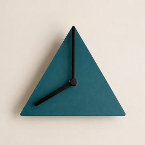 Könnte beinhalten: Eine dreieckige Wanduhr mit einem matten, blaugrünen Zifferblatt und drei dünnen schwarzen Zeigern. Die Uhr hat ein schlichtes, geometrisches Design ohne Zahlen oder Markierungen auf dem Zifferblatt, die Zeiger stehen auf etwa 7:40 Uhr.