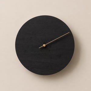 May include: A circular wall clock featuring a solid matte black wooden face with a visible wood grain texture. The clock has a single thin, metallic gold-toned hand attached to a central hexagonal gold-toned hub, indicating the time.