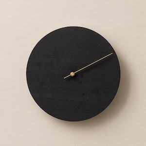 Könnte beinhalten: Eine kreisförmige Wanduhr mit einem massiven, mattschwarzen Holzgehäuse, bei dem die Maserung des Holzes sichtbar ist. Die Uhr verfügt über einen einzelnen, dünnen, goldfarbenen Zeiger, der an einer zentralen, sechseckigen Nabe befestigt ist.