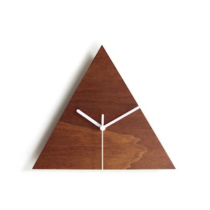 Puede incluir: Un reloj de pared moderno y minimalista con forma de triángulo, hecho de madera marrón oscura. Las manecillas y la línea que indica la hora son blancas. Un elemento decorativo para cualquier habitación.