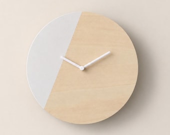 May include: A circular wall clock featuring a split design with one side in natural light wood grain and the other side in a solid matte white finish. The clock includes two simple white hands centered on the face, with no visible numbers or markers.