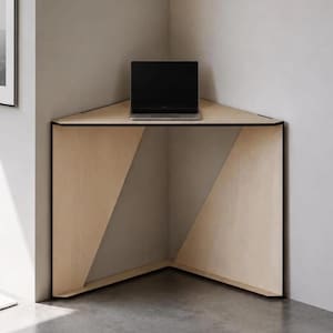 May include: A light-coloured wooden corner desk with a triangular top and two solid side panels that meet at a right angle. The desk features a smooth, flat surface with a dark edge finish, and a black laptop is placed on the centre of the desk.