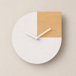 May include: A circular wall clock featuring a white finish with a square tan section in the upper right quadrant. The clock has two simple white hands centered on the face, with a flat right edge that interrupts the circular shape of the clock.