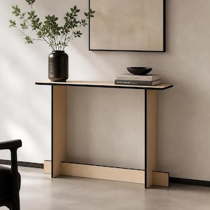 May include: A modern, minimalist console table with a light wood top and black trim. The table has a rectangular shape and is supported by four vertical legs. A black vase with greenery, books, and a black bowl are on the table.