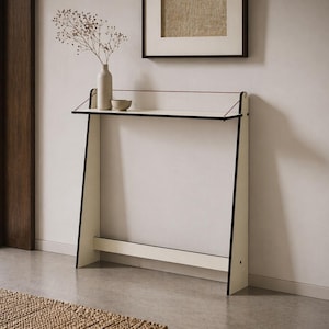 May include: A modern, cream-colored console table with black trim. The table has a rectangular top and angled legs. A small vase with dried flowers and a small bowl sit on the table. The table is placed against a white wall, with a framed picture above it.
