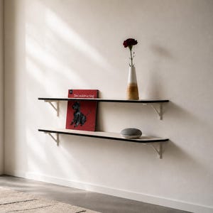 May include: Two horizontal wall-mounted shelves with white surfaces and black edges, supported by white metal brackets. The top shelf holds a red book titled Deconstructing and a tall vase with a dark flower. The bottom shelf holds a grey orb.