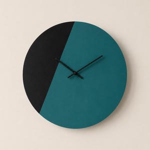 May include: A circular wall clock featuring a split design with one side in matte black and the other in deep teal. The clock face is devoid of numbers or markers and includes two thin, black hands positioned at the center to indicate time.