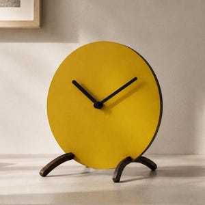 May include: A circular table clock with a bright yellow face and two thin black hands. The clock is supported by two dark brown wooden legs that curve outward, providing a stable base for the round timepiece on a flat surface.