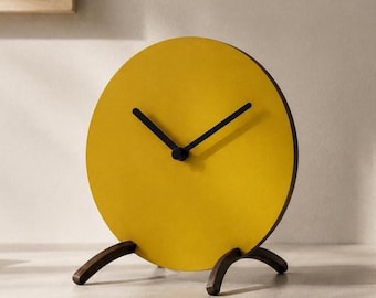 May include: A circular table clock with a bright yellow face and two thin black hands. The clock is supported by two dark brown wooden legs that curve outward, providing a stable base for the round timepiece on a flat surface.