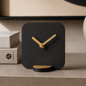 Puede incluir: Un reloj analógico cuadrado de color negro con manecillas doradas y una base semicircular a juego en color oro. La esfera del reloj es lisa, sin números ni marcas, con un acabado mate suave en el panel cuadrado y un soporte dorado.