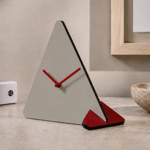 Puede incluir: Un reloj moderno y geométrico con una esfera triangular en gris claro, bordes negros y manecillas rojas. Una base roja y negra sostiene el reloj. El reloj es un elemento decorativo para un escritorio o estante.
