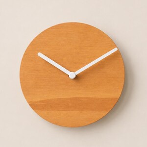 Può includere: Un orologio da parete circolare con un quadrante in legno liscio color marrone chiaro e due semplici lancette bianche centrate al centro. L'orologio è privo di numeri o indicatori delle ore, con una superficie in legno a venature.