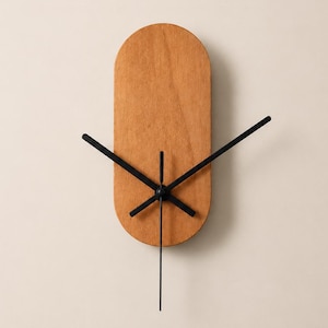 May include: A wall clock featuring a vertical, pill-shaped wooden face with a natural light brown grain. The clock has three thin, black metal hands positioned at the center, extending outward to indicate the time against the wooden surface.