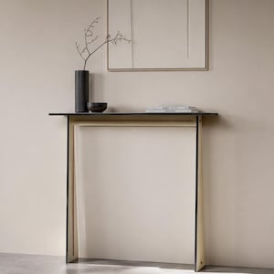 May include: A contemporary console table with a black surface and a light-toned frame. A black vase with a sprig of branches, a small black bowl, and books are on the table. The table is against a neutral wall.