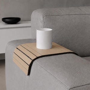 Op de afbeelding: Een lichtbeige houten sofa-armtafel met zwarte rand, met een witte cilindrische mok. De tafel heeft een lattenontwerp en is geplaatst op een grijze leren sofa-arm. Een zwarte kom staat op een witte kast op de achtergrond.