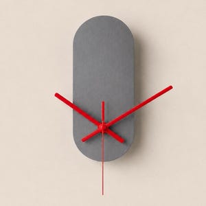 May include: A wall clock featuring a vertical, pill-shaped grey face with rounded ends. The clock has three bright red hands positioned at the centre, with the hour, minute, and second hands extending outward against the solid grey background.
