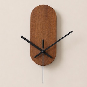 May include: A wall clock featuring a vertical, pill-shaped wooden face with a medium brown wood grain finish. The clock has three thin, black metal hands positioned at the centre, extending outward to indicate the time without any numerical markings.