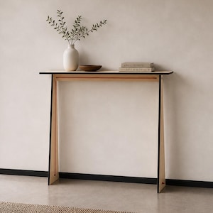 Peut inclure: Une console moderne avec un plateau noir et des pieds en bois clair. Un vase blanc avec de la verdure, un petit bol en bois et deux livres sont exposés sur la table. La table est placée contre un mur neutre.