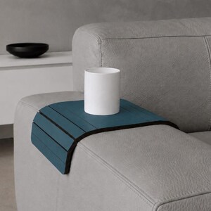 May include: A teal blue sofa arm tray with a black trim, holding a white cylindrical mug. The tray is made of wooden slats. The sofa is gray and has a modern design. A black bowl sits on a white cabinet in the background.