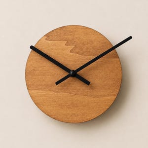 Könnte beinhalten: Eine kreisförmige Wanduhr mit einem Zifferblatt aus naturfarbenem, hellbraunem Holz mit sichtbarer Maserung und glatter Oberfläche. Die Uhr hat drei dünne, gerade Zeiger aus schwarzem Metall in der Mitte, ohne Ziffern oder Markierungen.