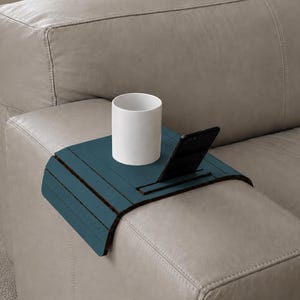 Puede incluir: Una bandeja de brazo de sofá de madera azul verdoso con una ranura para un teléfono y una taza de cerámica blanca. La bandeja se coloca en un brazo de sofá de cuero gris claro. La bandeja está diseñada para sostener bebidas y dispositivos electrónicos.