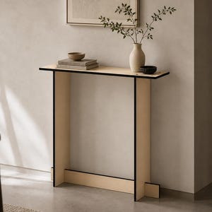 May include: A modern, minimalist console table with a light wood finish and black trim. The table features a rectangular top and two supporting legs. A vase with greenery, a small bowl, and books are displayed on the table.
