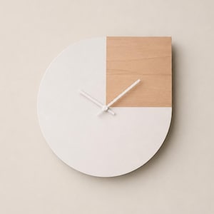 May include: A circular wall clock featuring a matte white finish across most of its surface, with a square section of light-toned natural wood grain in the upper right quadrant. The clock has two simple white hands centered on the face.