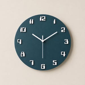 May include: A circular wall clock with a dark teal face and white, block-style numerals from one to twelve. The clock features three thin, white hands positioned at the center, indicating the time against the solid, matte-finish background.