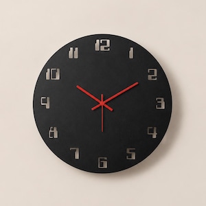 May include: A circular wall clock with a matte black face featuring cut-out silver-toned numerals from one to twelve. The clock has three thin, bright red hands for the hour, minute, and second indicators, positioned centrally on the dark surface.