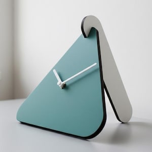 May include: A contemporary teal and white triangular desk clock. The clock face is teal with white hands and a silver centre. The clock has a white stand and a black border. A minimalist design.