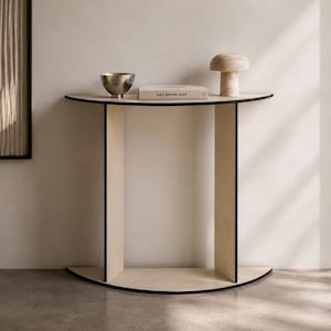 Puede incluir: Una mesa consola semicircular hecha de madera clara con bordes negros. Sobre la superficie hay un cuenco metálico, un libro titulado EAT DRINK NAP y una lámpara de madera con forma de seta. La mesa cuenta con dos soportes verticales.