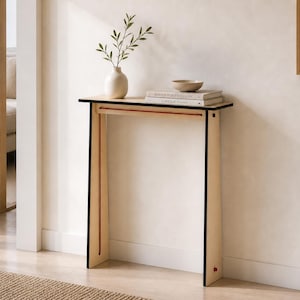 May include: A light-coloured wooden console table with black edges and a thin red accent line along the top frame. The table features a flat rectangular top supported by two vertical side panels, holding a white vase with greenery and two books.
