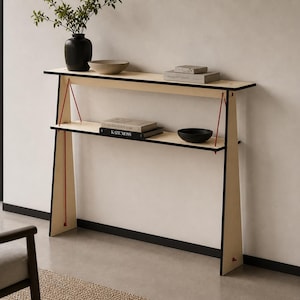May include: A contemporary console table in light wood with black edging and red support cords. It features two shelves holding a black vase with foliage, a beige bowl, books, and a black bowl. The design is minimalist and stylish.