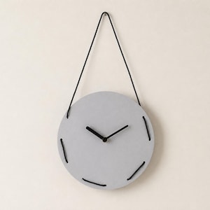 Könnte beinhalten: Eine kreisförmige, hellgraue Wanduhr mit glatter, matter Oberfläche. Das Zifferblatt hat schwarze Zeiger und wird von einer schwarzen Kordel gehalten, die durch den Rand des runden Gehäuses geführt ist, was eine einfache Zeitmessung ermöglicht.