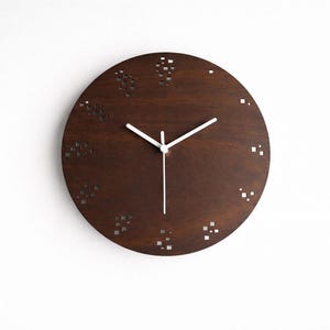 May include: A round, dark brown wooden wall clock with white hands. The clock face features small, square cutouts arranged in clusters. The clock is mounted on a white wall, creating a minimalist aesthetic.
