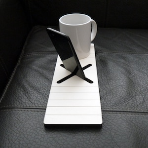 Puede incluir: Soporte de teléfono y taza de madera blanca con un teléfono inteligente negro y una taza blanca en la parte superior. El soporte tiene un recorte en forma de cruz en el medio.