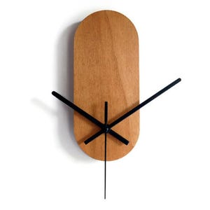 Puede incluir: Un reloj de pared minimalista con una esfera de madera de color marrón claro y forma ovalada. El reloj presenta manecillas negras y un segundero negro. El diseño es simple y moderno, adecuado para la decoración del hogar.